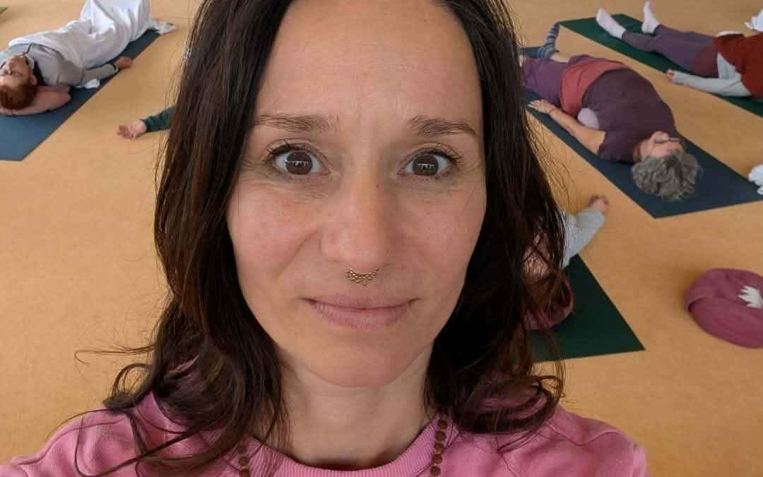 Julia lächelt bei einem Selfi in die Kamera im Hintergrund ist die Gruppe Yogalehrer in einer Endentspannung